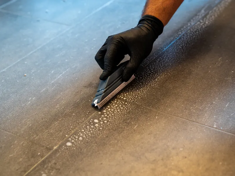 a professional cleaner cleaning grout with a brush blade and foamy soap on a gray tiled bathroom floor